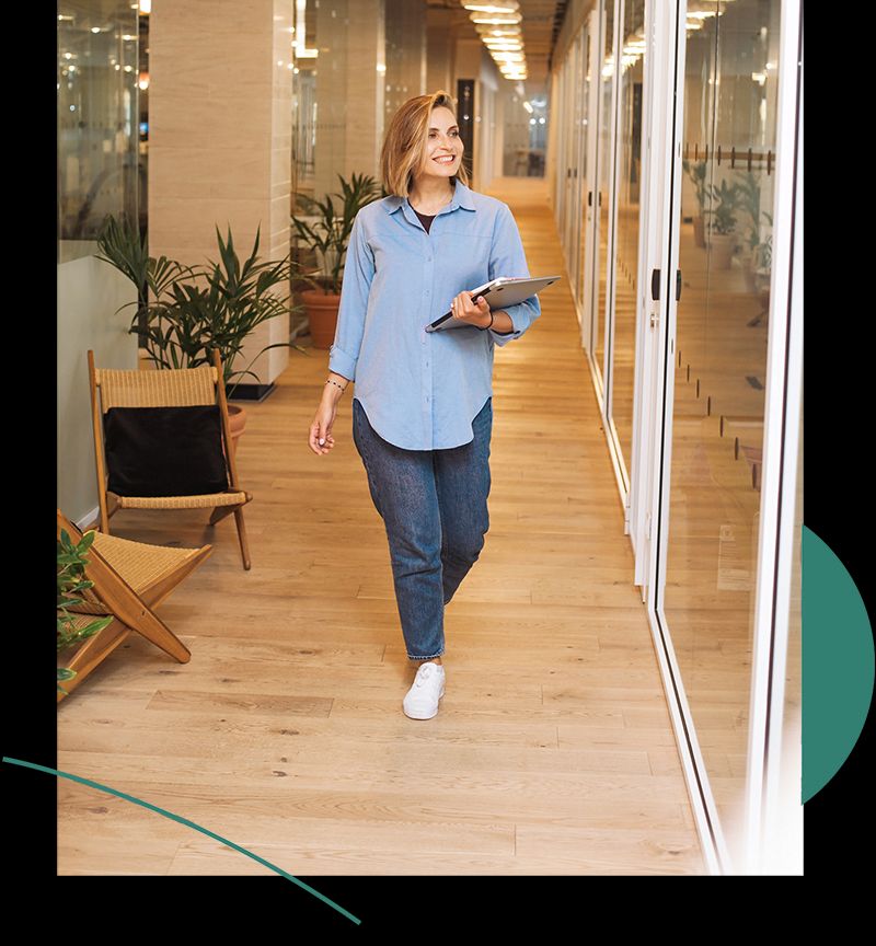 A woman walks down an office hallway, she is holding a laptop in her left hand, smiling.
