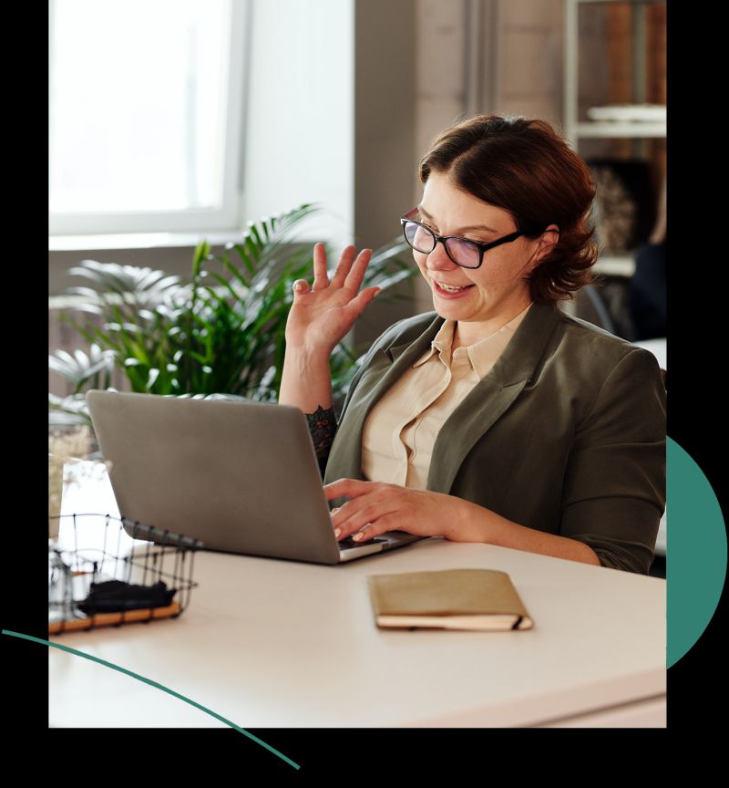 A woman sitting at a desk while using a laptop. She is smiling and waving towards the laptop, indicating an ongoing video meeting.